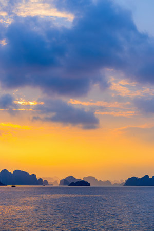 A stunning view of Ha Long Bay at sunset, with the sky illuminated in warm golden hues. Dark limestone islands and cliffs are silhouetted against the serene water and glowing horizonの写真素材
