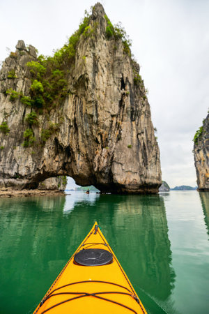 A kayak approaches a natural limestone arch in Ha Long Bay. Towering cliffs and calm, green waters create a peaceful yet adventurous setting in this world-renowned Vietnamese destinationの写真素材