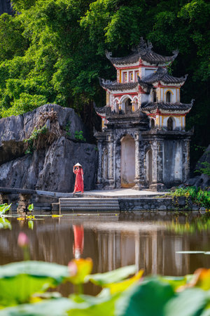A woman in a red dress and conical hat stands near the iconic Bich Dong Pagoda, surrounded by lush greenery and a lotus-filled pond in Tam Coc, Vietnamの写真素材