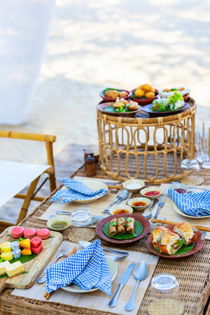 A scenic picnic arrangement on a beach featuring a wicker table set with traditional Vietnamese snacks, fruits, spring rolls, banh mi sandwiches, and a variety of sauces, with a wicker serving stand nearbyの写真素材