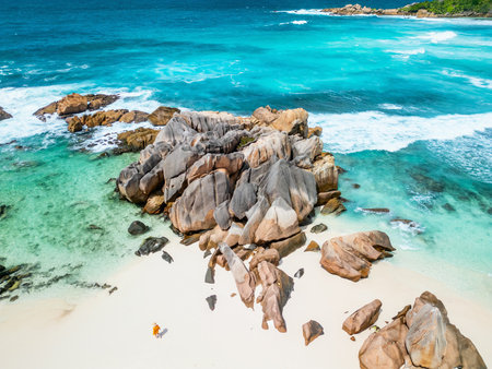 An aerial view captures a person in a yellow outfit standing on the white sandy beach of La Digue, Seychelles, surrounded by striking granite rocks and turquoise watersの写真素材