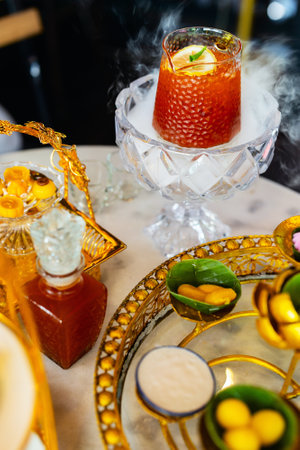 A decorative setup featuring a glass of iced lemon tea on dry ice, creating a smoky effect, surrounded by a variety of traditional desserts and ornate golden serving traysの写真素材