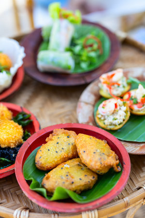 An assortment of traditional Vietnamese snacks including fried banana fritters, spring rolls, and shrimp-topped rice cakes, all served on colorful plates with banana leaf accentsの写真素材
