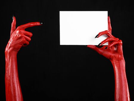 Halloween theme: Red devil hand with black nails holding a blank white card on a black background studioの写真素材