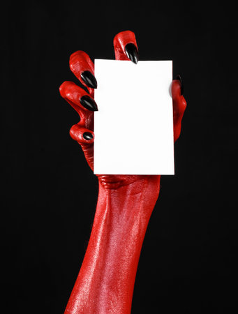 Halloween theme: Red devil hand with black nails holding a blank white card on a black background...