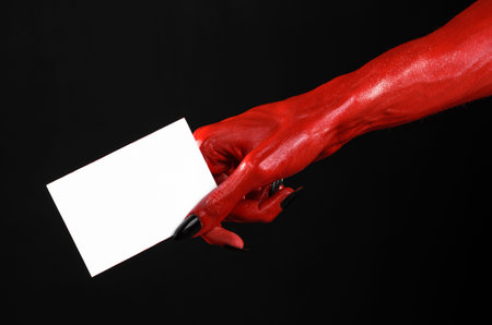 Halloween theme: Red devil hand with black nails holding a blank white card on a black background...