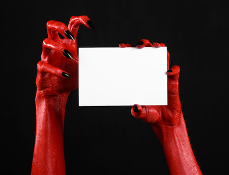 Halloween theme: Red devil hand with black nails holding a blank white card on a black background...