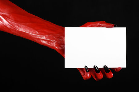 Halloween theme: Red devil hand with black nails holding a blank white card on a black background...
