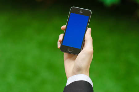 Communication and Business Subject: Hand in a black suit holding a modern phone with blue screen in the background of green grass studioの写真素材