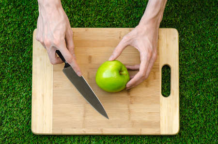 Vegetarians and cooking on the nature of the theme: human hand holding a knife and green apple on the background of a cutting board and green grassの写真素材