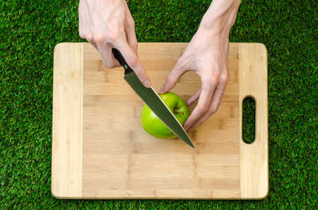Vegetarians and cooking on the nature of the theme: human hand holding a knife and green apple on the background of a cutting board and green grassの写真素材