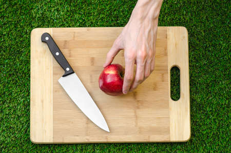 Vegetarians and cooking on the nature of the theme: human hand holding a knife and a red apple on the background of a cutting board and green grassの写真素材