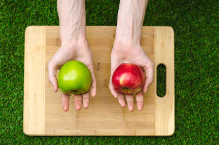 Vegetarians and cooking on the nature of the theme: human hand holding red and green apples on the background of a cutting board and green grassの写真素材