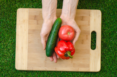 Vegetarians and cooking on the nature of the theme: human hand holding cucumber, tomato and red pepper on a cutting board and a background of green grassの写真素材