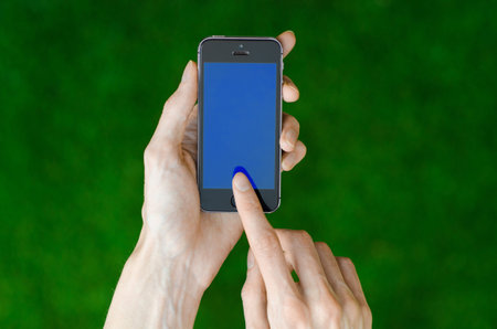 Human hand holding a modern mobile phone with a blue screen on a background of green grass...