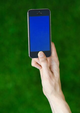 Human hand holding a modern mobile phone with a blue screen on a background of green grass first-person view studioの写真素材