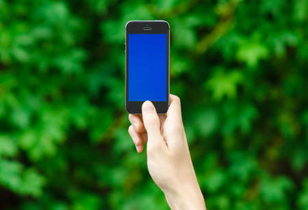 Human hand holding a modern mobile phone with a blue screen on a background of green grass first-person view studioの写真素材