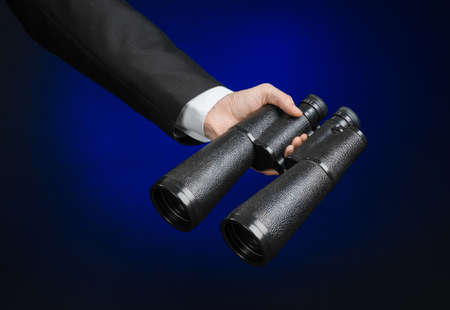 Business and search topic: Man in black suit holding a black binoculars in hand on a dark blue background in studioの写真素材
