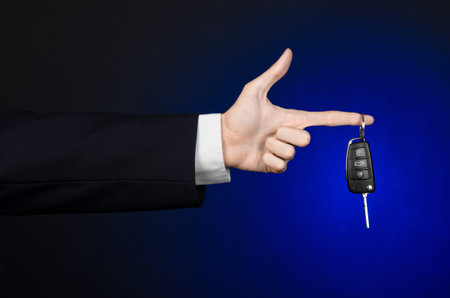Business and gift theme: car salesman in a black suit holds the keys to a new car on a dark blue background in studioの写真素材