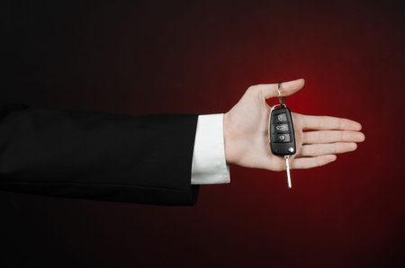 Business and gift theme: car salesman in a black suit holds the keys to a new car on a dark red...