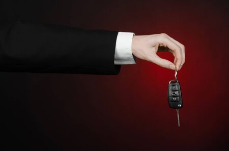 Business and gift theme: car salesman in a black suit holds the keys to a new car on a dark red background in studioの写真素材
