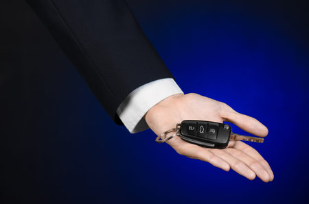 Business and gift theme: car salesman in a black suit holds the keys to a new car on a dark blue...