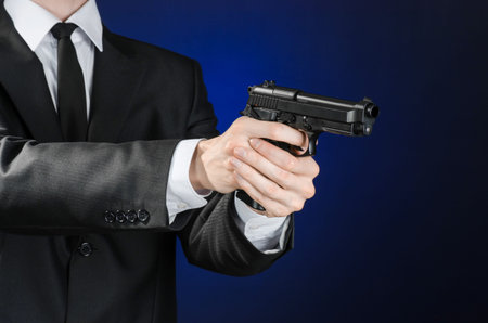 Firearms and security topic: a man in a black suit holding a gun on a dark blue background in studioの写真素材