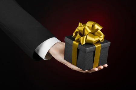 The theme of celebrations and gifts: a man in a black suit holding a exclusive gift packaged in a black box with gold ribbon, beautiful and expensive gift on a dark red backgroundの写真素材