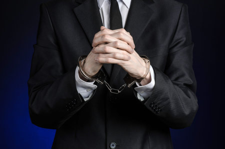 Corruption and bribery theme: businessman in a black suit with handcuffs on his hands on a dark blue background isolatedの写真素材
