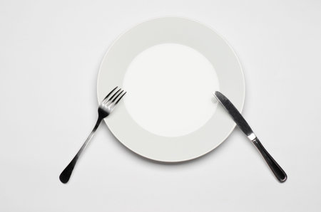 Cutlery and restaurant topic: Fork knife and white plate lying on a white table isolated top view