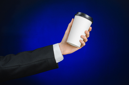 Business lunch and coffee theme: businessman in a black suit holding a white blank paper cup of coffee with a brown plastic cap on a dark blue background isolated in the studioの写真素材