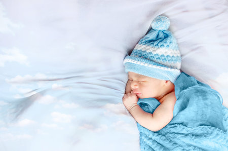 Newborn baby boy sleeping on white silk bed floating clouds wearing blue hat and blanket isolated in...