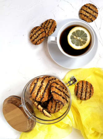 cookies polished with chocolate and peanut pieces in a glass jar with a wooden lid on a light table a Cup of tea with a slice of lemon and a yellow runner.の写真素材