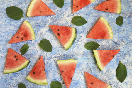 a circle of sliced triangles of watermelon with green mint leaves on a blue solid background , the concept of minimalismの写真素材