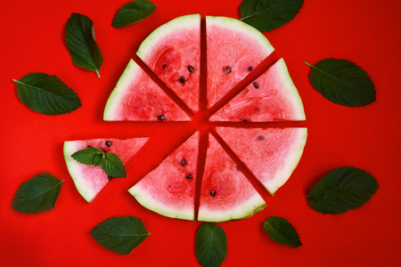 a circle of sliced triangles of watermelon with green mint leaves on a red solid background , the concept of minimalismの写真素材