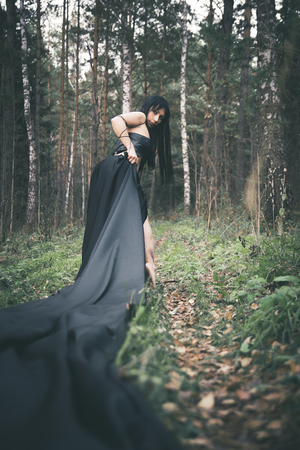 girl witch brunette in black leather top and long black skirt with long black hair in autumn forest on all saints day, concept-Halloweenの写真素材