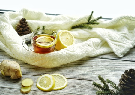 Glass cup of tea with lemon and ginger root slices, mint leaf on glass saucer on a rustic wooden table on background of the women's woolen mittens and knitted scarf outdoor.の写真素材