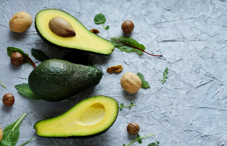 Avocado cut in half lies on a gray concrete background surrounded by nuts and green salad leaves...