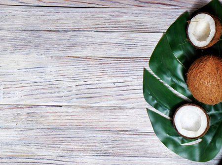two halves and one whole coconut with a leaf of tropical monstera plant on a white wooden...