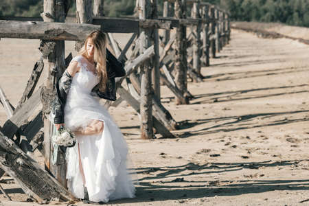 Blonde girl in a wedding dress and a leather jacket in a rock style on a wild sandy beach. high quality photoの写真素材