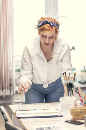 A young active female artist, with red hair, in a white shirt and jeans with a bandage on her head, paints standing at the table. high quality photoの写真素材