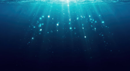 Sunlight rays pierce the dark blue ocean depths, illuminating tiny particles and creating a serene underwater scene.の素材