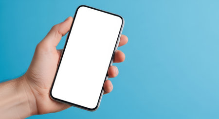 A person's hand holds a modern smartphone with a blank, white screen against a solid blue background. The phone is tilted slightly.の素材