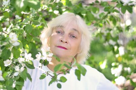 Beautiful older woman with blond hair next to a blooming tree in the garden in mayの写真素材
