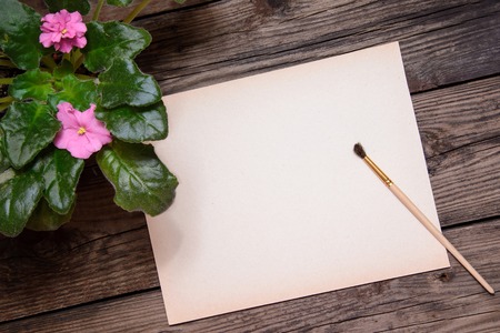 A pot of pink violet and green leaves, a piece of paper and a brush to paint on a brown wooden background with copy spaceの写真素材
