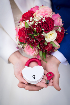 White with red wedding lock and key and a wedding bouquet of red and pink roses in hands of newlyweds close upの写真素材