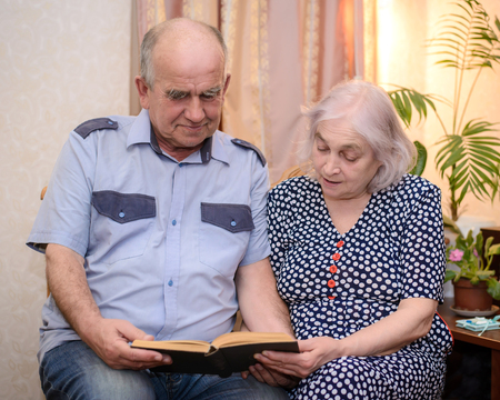 An elderly couple - grandfather in the blue shirt and the grandmother in the blue dress with white polka dots holding a book and reading it together at homeの写真素材