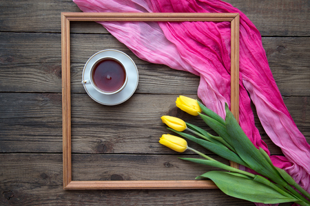 Beautiful romantic picture with frame and yellow tulips, pink cloth and a cup of tea on wooden background, top view with copy spaceの写真素材