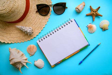 The layout on the theme of holidays at sea: straw hat, sunglasses, seashells and notepad with pen on a blue background, top viewの写真素材