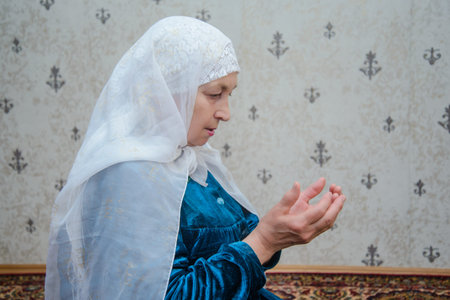 An elderly Muslim woman in a white scarf and a blue dress praying in the room